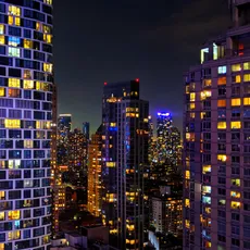 A vibrant night cityscape in Manhattan, showcasing illuminated skyscrapers with colorful windows and bright lights against a dark sky. The buildings create a mosaic of urban life and energy.