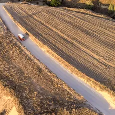 Aerial view of a red and white van driving along a narrow dirt road surrounded by dry, golden fields in Cappadocia. The landscape features patches of grass, trees, and lines from tire tracks. Shadows from the morning light are visible.