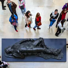 A group of people observes a sculpture, Kehinde Wiley’s reinterpretation of “Woman Bitten by a Serpent”, displayed on a dark platform in Musée d'Orsay. The statue depicts a reclining figure. The floor is light-toned, and visitors are casually dressed, indicating a busy exhibition space.