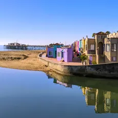 Colorful beachside apartments reflect in calm water at Venetian Court, Capitola. A sandy beach and pier stretch into the ocean under a clear blue sky.