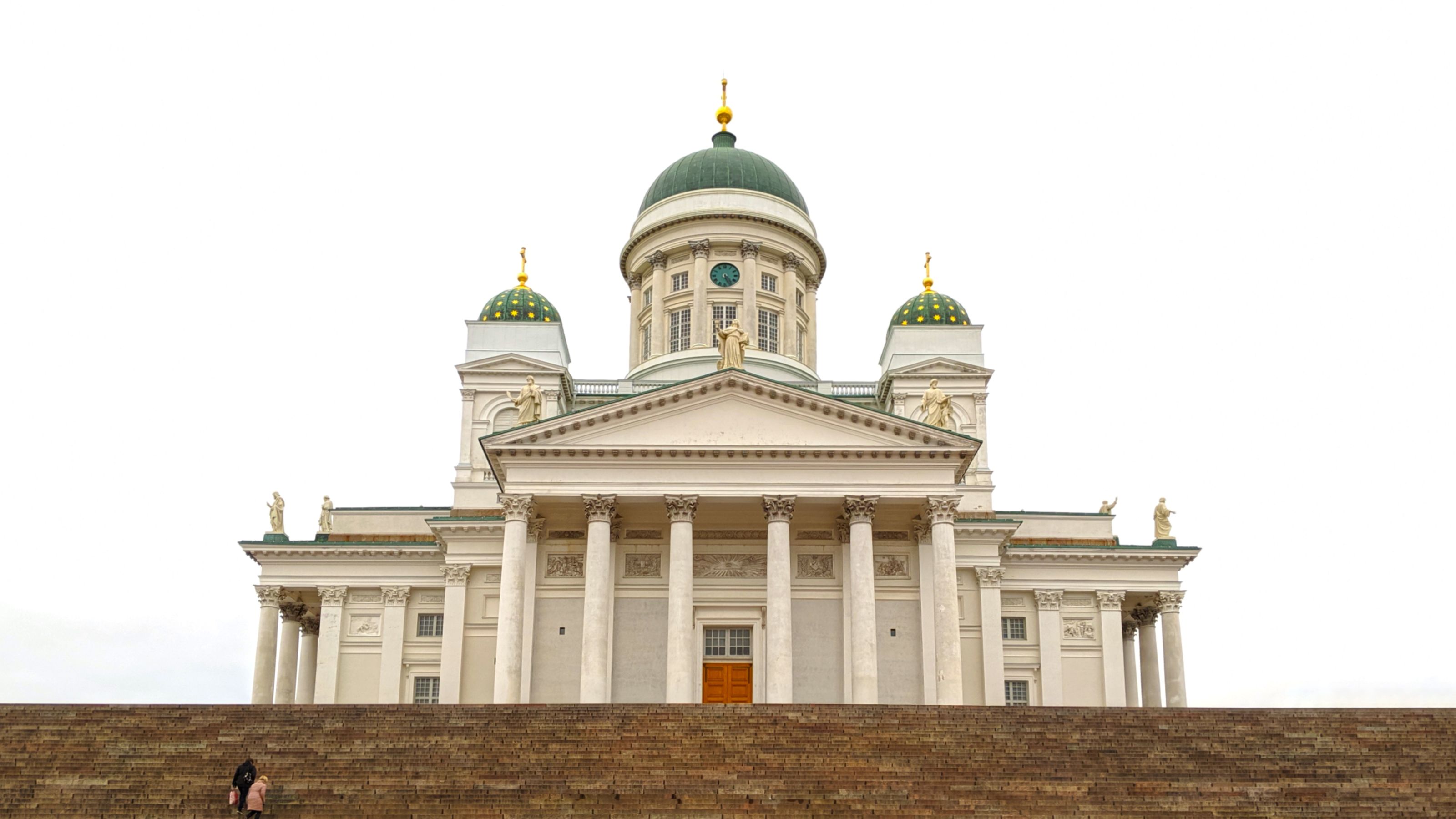 Helsinki Cathedral with its grand neoclassical facade and green domes under a bright white sky. The steps leading up are mostly empty, with two people near the bottom.