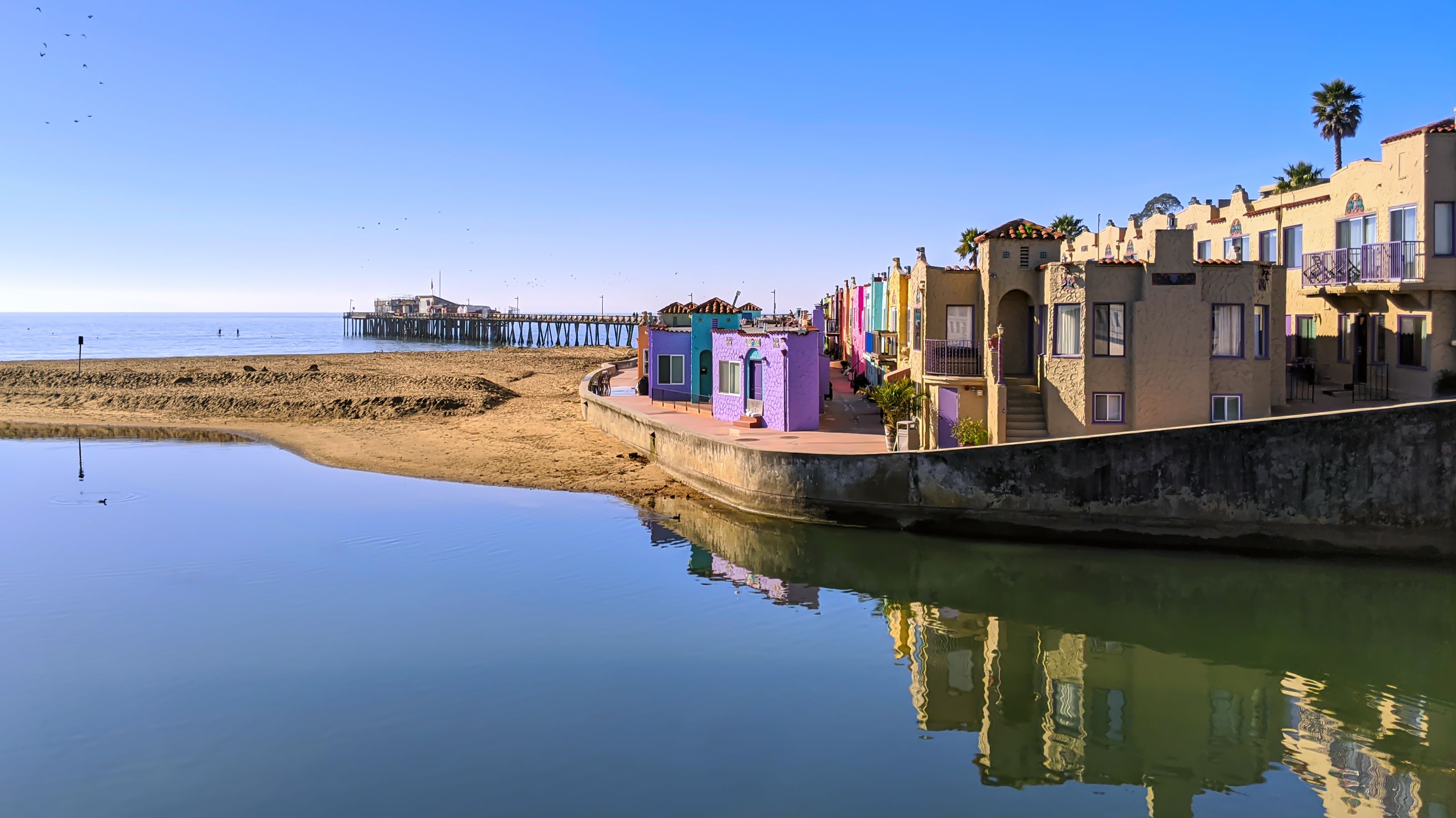 Colorful beachside apartments reflect in calm water at Venetian Court, Capitola. A sandy beach and pier stretch into the ocean under a clear blue sky.