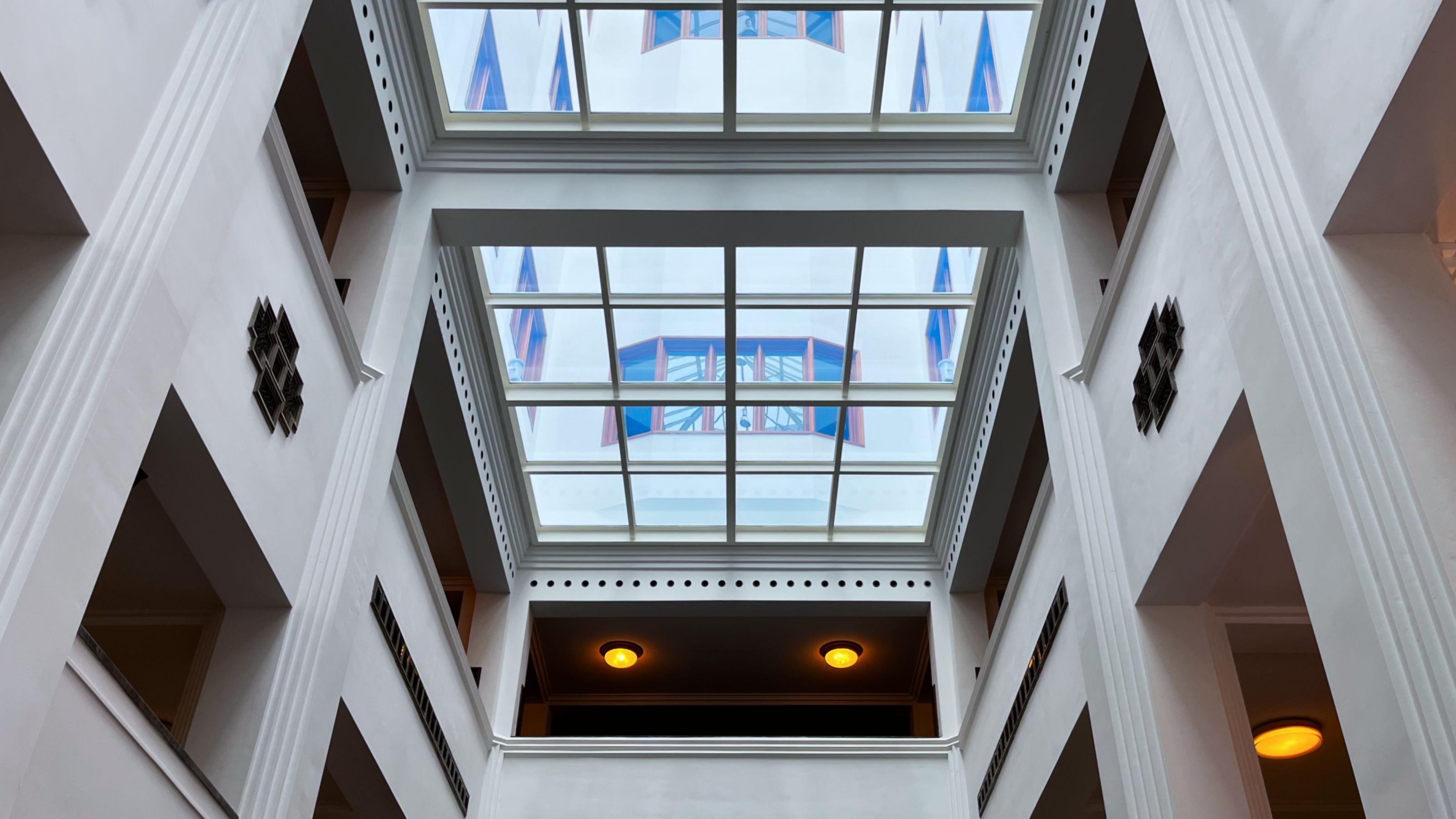 View of the Stadsarchief Amsterdam interior, featuring a grid skylight with geometric designs on white walls. The ceiling showcases colorful windows and lit ceiling lamps, creating an open and bright atmosphere.