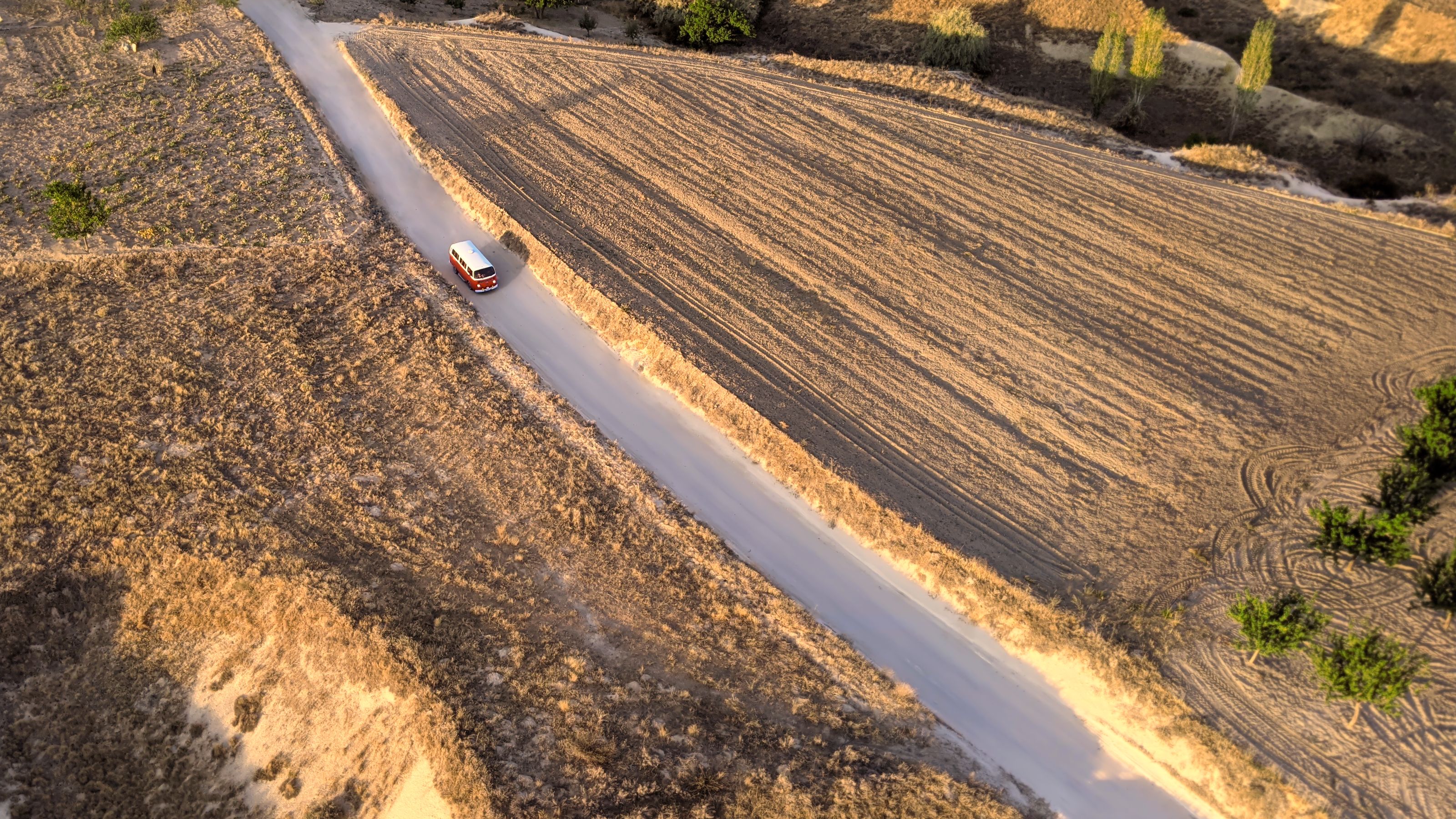 Aerial view of a red and white van driving along a narrow dirt road surrounded by dry, golden fields in Cappadocia. The landscape features patches of grass, trees, and lines from tire tracks. Shadows from the morning light are visible.