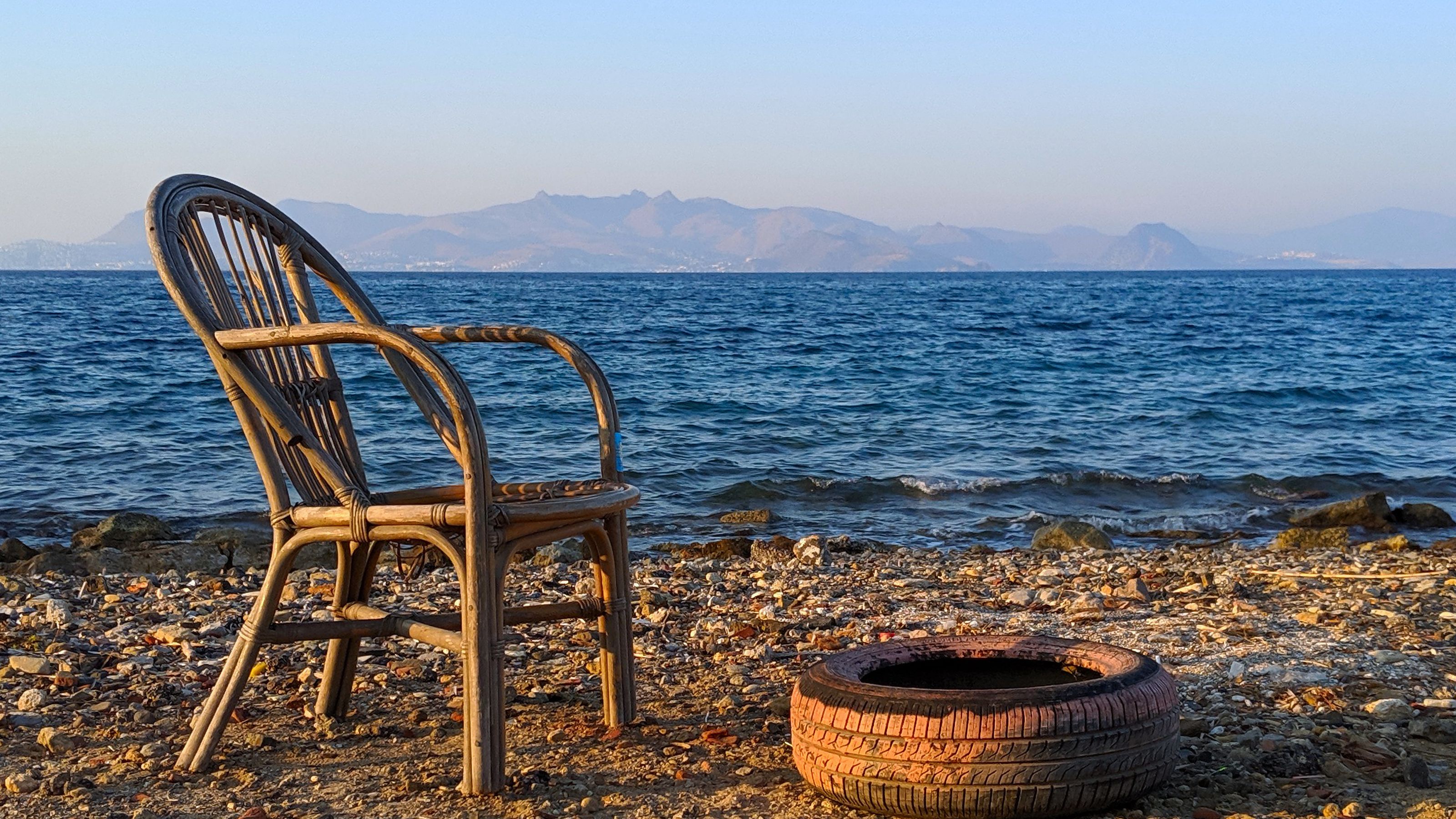 A wicker chair and a car tire on a pebbly beach, with the sea and distant mountains in the background.