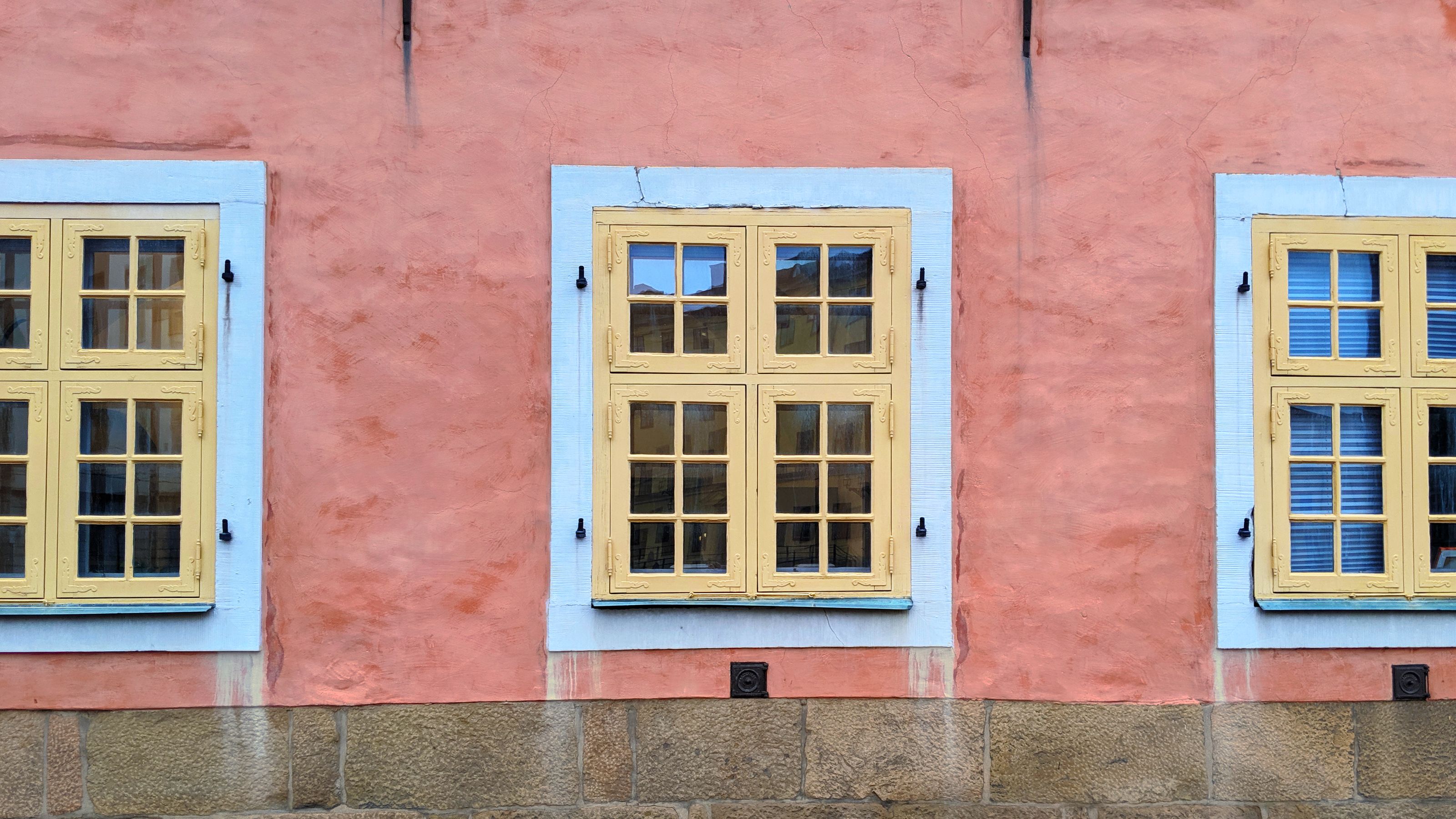 Three yellow-framed windows on a pink pastel wall with a stone foundation.