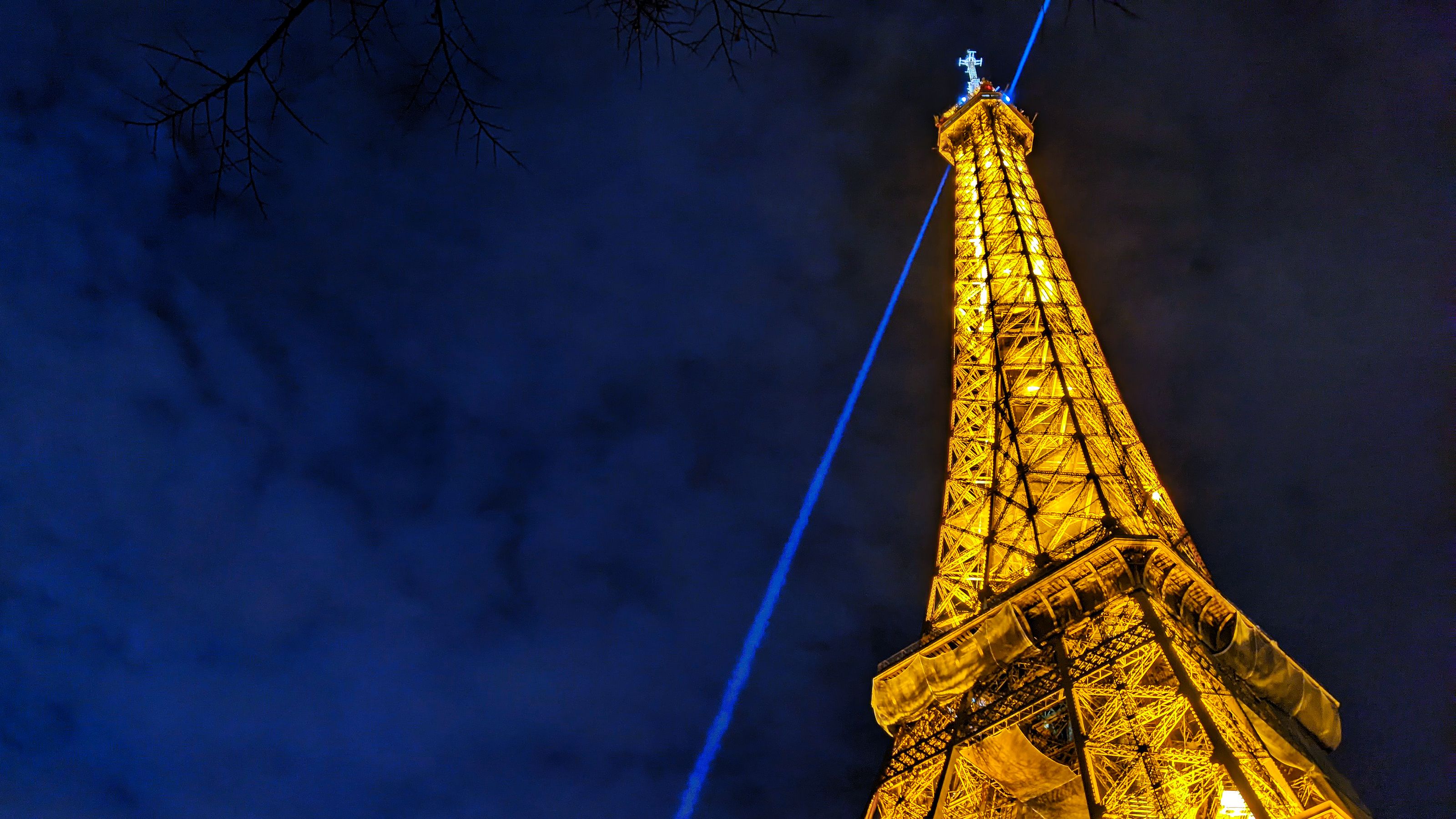 Illuminated Eiffel Tower at night, with a blue light beam from its peak against a dark blue sky, framed by faint tree branches.