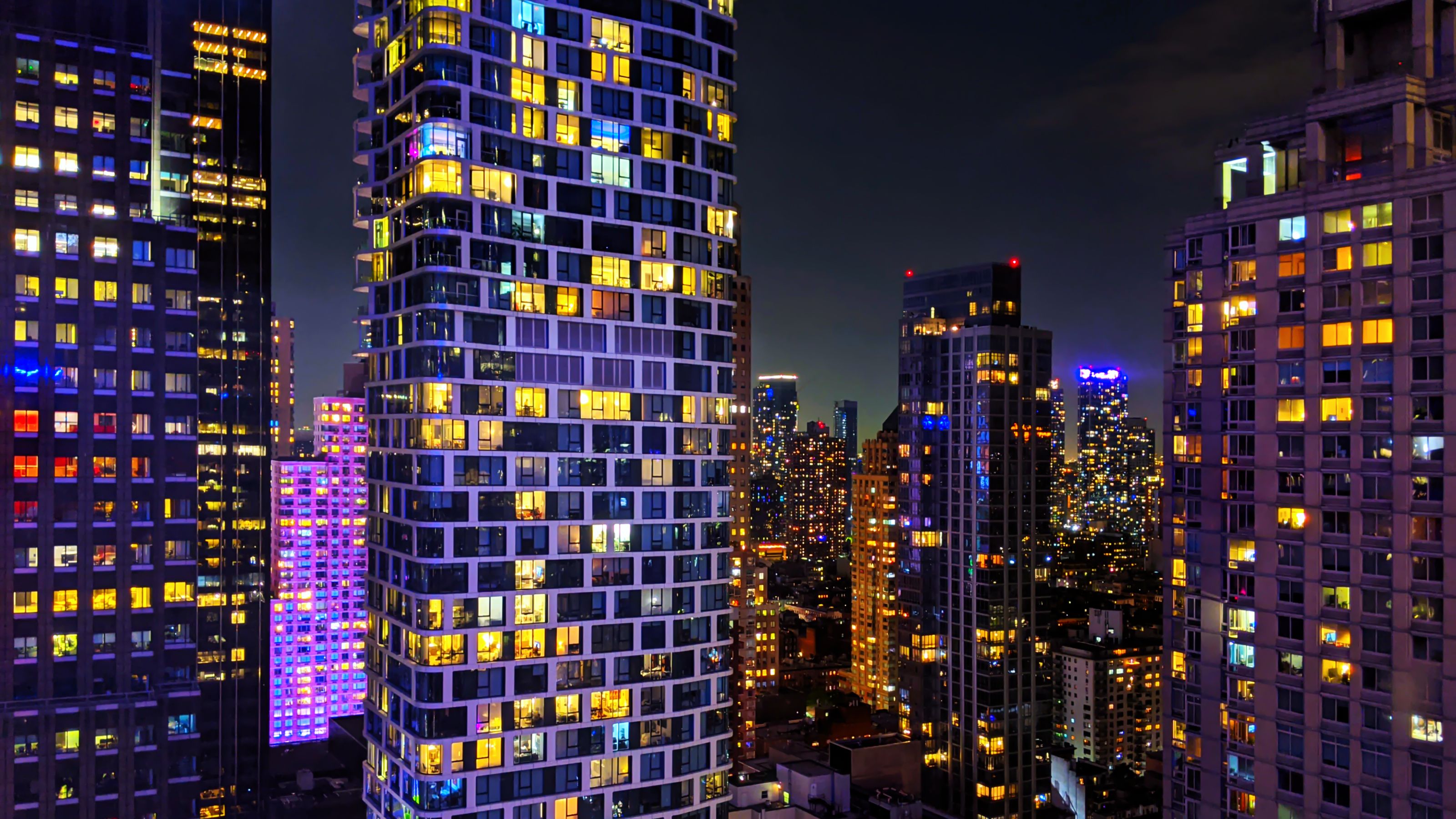 A vibrant night cityscape in Manhattan, showcasing illuminated skyscrapers with colorful windows and bright lights against a dark sky. The buildings create a mosaic of urban life and energy.