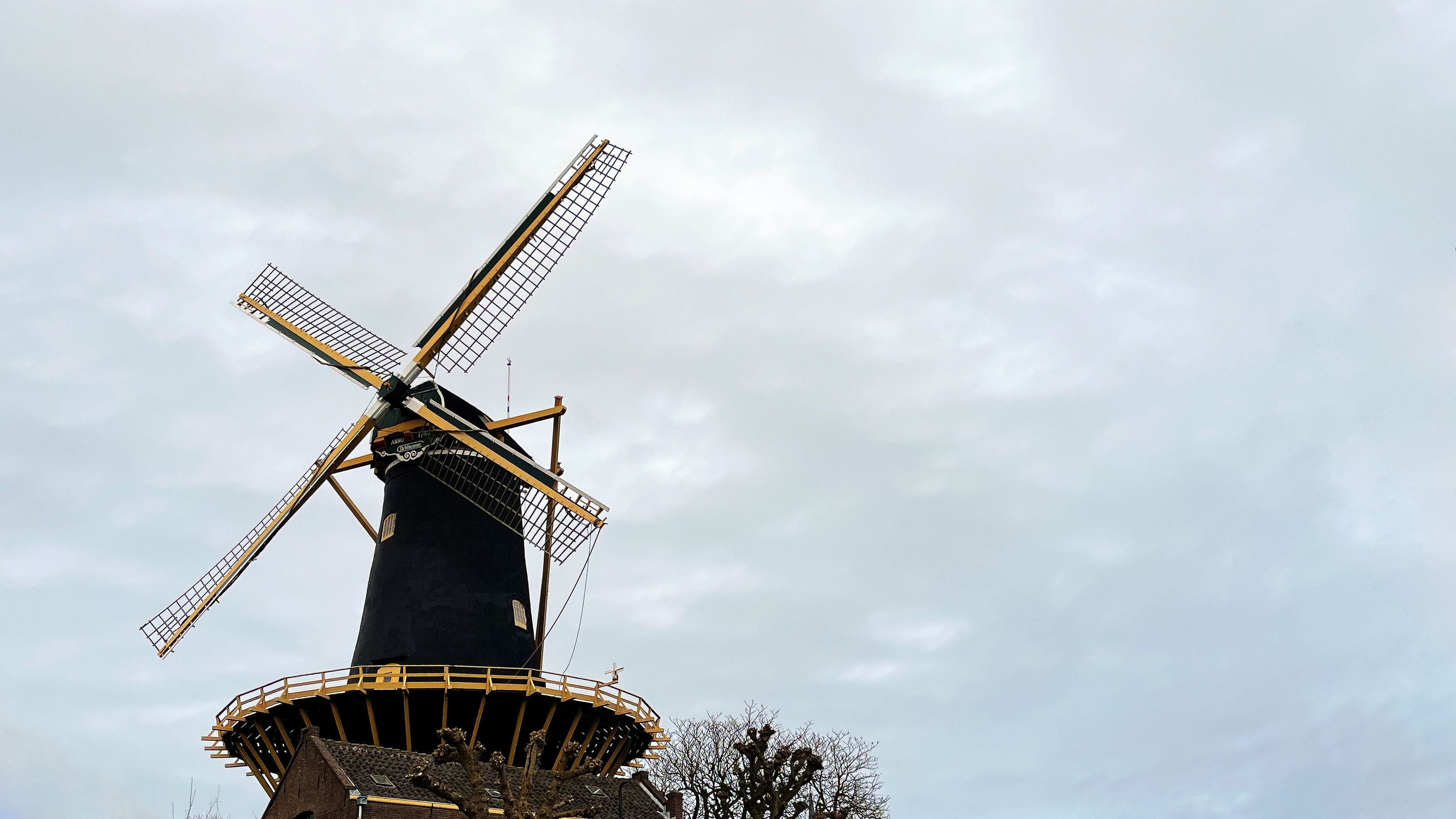 A traditional Dutch windmill with a black base, a band of decoration, and yellow blades against a cloudy sky in Woerden.