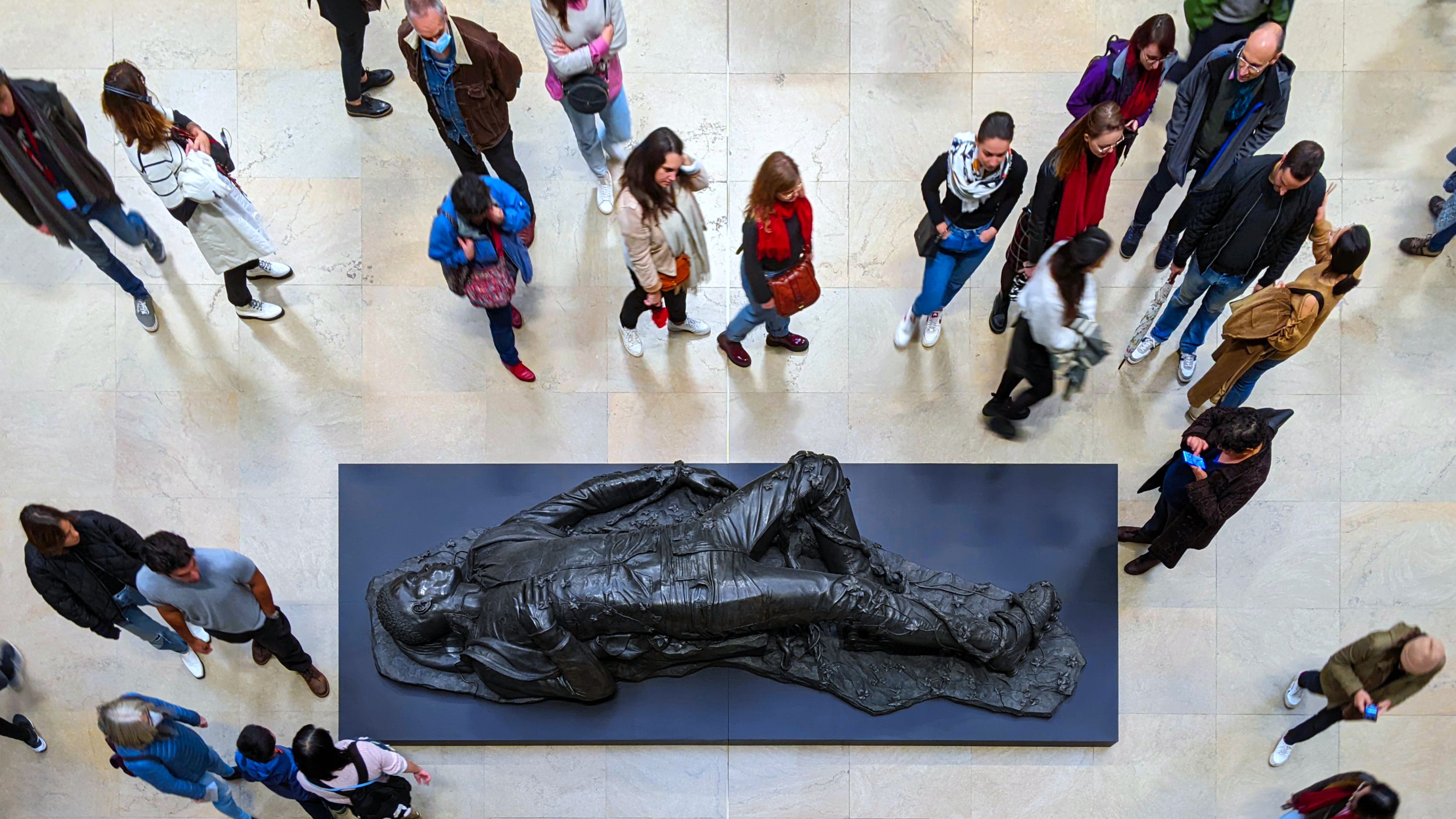 A group of people observes a sculpture, Kehinde Wiley’s reinterpretation of “Woman Bitten by a Serpent”, displayed on a dark platform in Musée d'Orsay. The statue depicts a reclining figure. The floor is light-toned, and visitors are casually dressed, indicating a busy exhibition space.